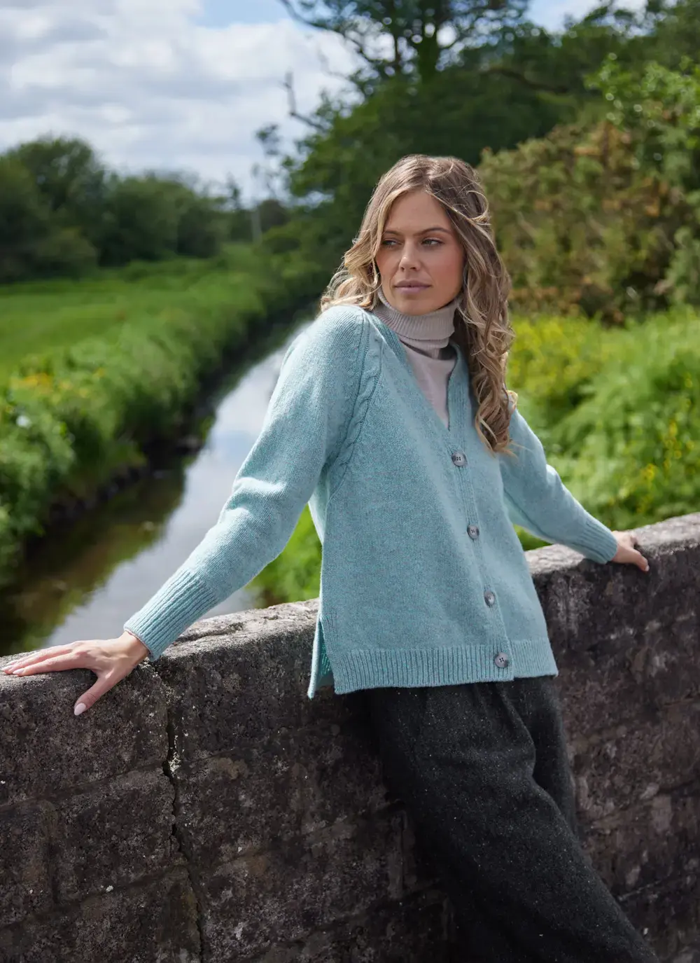Woman wearing a sea stone Fisherman cashmere raglan cable V-neck cardigan, styled over a beige turtleneck by a stone wall in the countryside.
